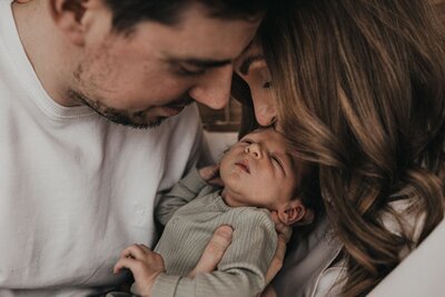 A close-up in soft monochrome of a newborn baby's tiny hand gripping their father's finger, while the mother lovingly looks on. Tender family momen