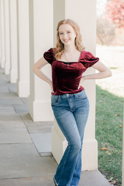 Senior girl in velvet burgundy shirt and jeans stands in front of a piller at the Lily House, Indianapolis senior photographer