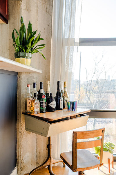 A small, vintage-style desk has been repurposed as a bar cart. Several bottles of liquor, including vodka, gin, and wine, are arranged on the wooden surface. A set of colorful shot glasses sits to the right. The desk has a pull-out drawer and is positioned next to a large window with sheer white curtains. A potted snake plant sits on a shelf above the desk against a textured concrete wall. A wooden chair with a padded seat is tucked under the desk.
