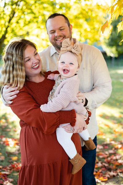 Parents hold their toddler and look at her smiling in the midst of fall foliage in Ann Arbor