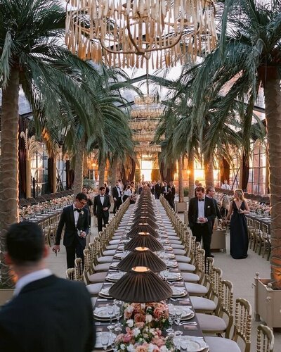 An elegant long dining table setup under a pavilion with palm trees and chandeliers.