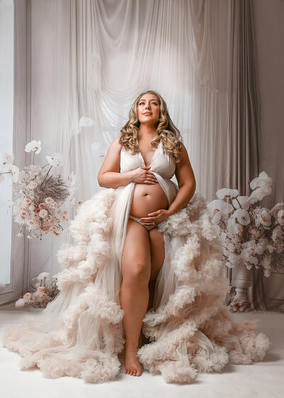 Pregnant mother with blonde hair, wearing a tulle gown looking up , against a neutral tan floral and curtain background in my Arizona Maternity studio