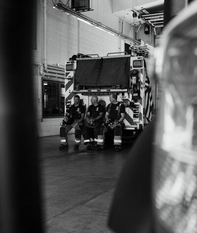 Black and white scene of fire fighters sitting on the back of their firetruck