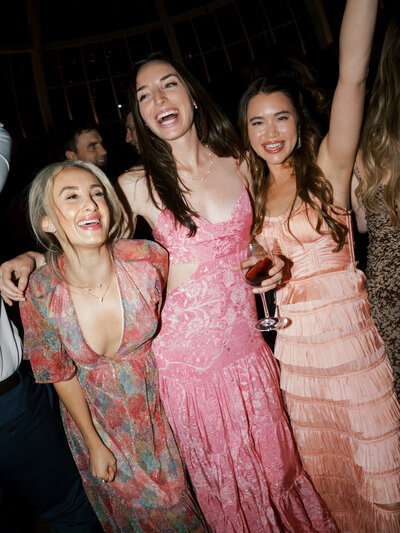 Guests dancing on the dance floor at the wedding reception with their arms in the air and colorful dresses on.