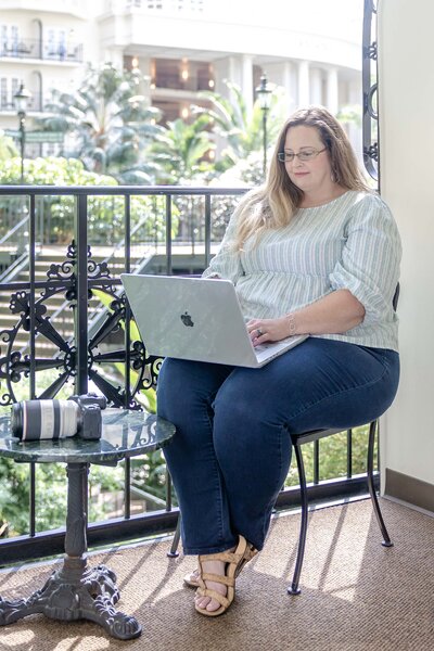 A woman with long, straight hair sits comfortably at a small table, focused on her laptop. She wears a light, patterned top and dark jeans, with a camera and lens beside her, suggesting she is a photographer planning a North Carolina wedding shoot. The bright, airy space features lush greenery in the background.