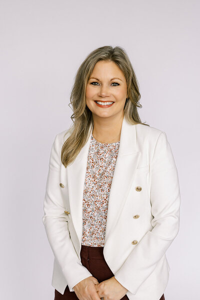 Smiling woman with long hair, wearing a white blazer over a floral blouse, in front of a plain background.