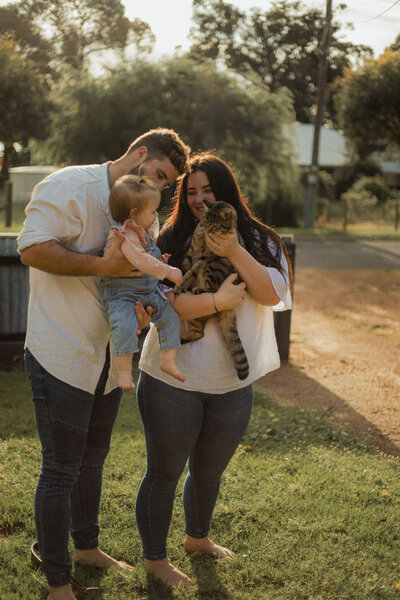 Couple standing close together, his arms around her waist, her hand on his chest in front of greenery and backlit with golden light