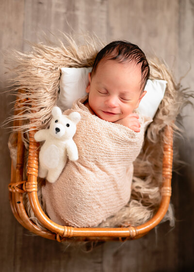 Baby boy wrapped in tan swaddle blanket sleeps in basket with white teddy bear.
