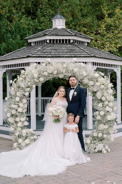 Bride and groom with flower girl in front of floral arch at Deer Creek wedding venue in Ajax, photographed by a Toronto-based wedding photographer