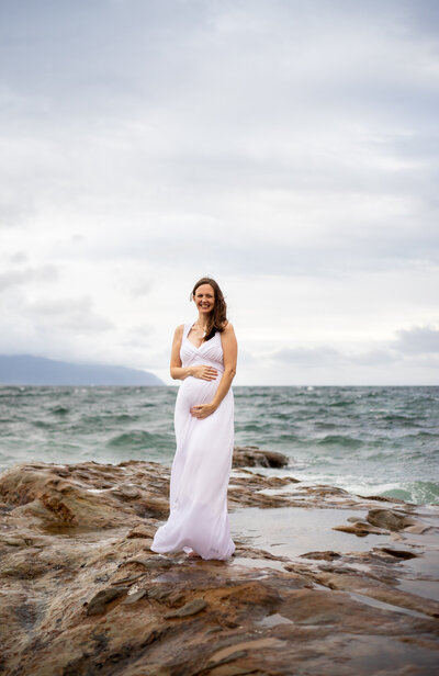 Pregnant woman in white long maxi dress standing on the rock of Cape Kiwanda , Oregon by Jaime Bugbee Photography