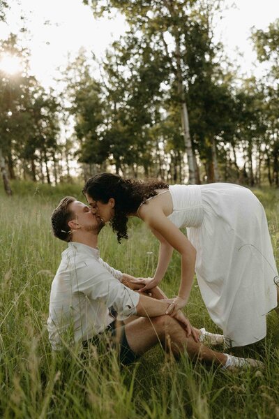 Engagement shoot of a couple in green grass, with the groom-to-be seated and his fiancée bending down to kiss him