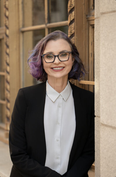 woman smiling at the camera leaning against an art deco window wearing glasses in a suit