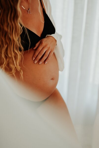 Close up image of a pregnant woman's belly. She is in a black bikini and stands near a window in a lovely photo studio