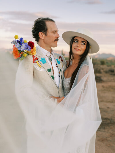 couple in desert embracing the bride is wearing a hat and the veil wraps around them