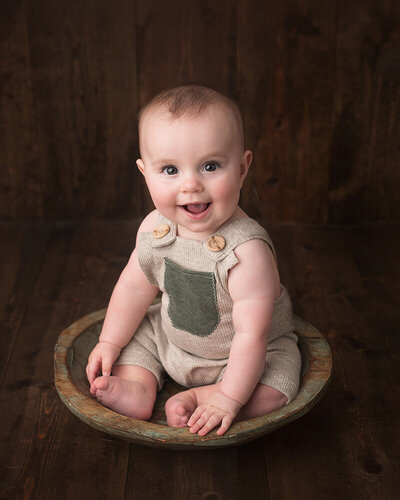 Studio baby milestone session photo of a six month old boy smiling, seated in a wooden bowl