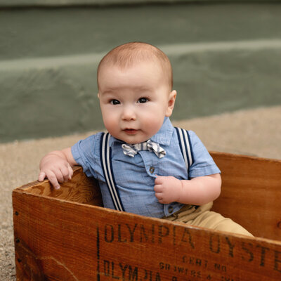 A little boy wearing a bowtie, suspenders, and a blue shirt sits in a crate