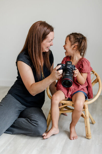 Melissa kneels next to her young daughter, both smiling and laughing while holding a camera together in a bright, minimal studio setting.