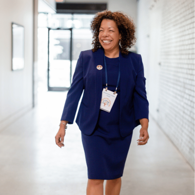 Photo of Michelle McKown-Campbell smiling and walking down a hall in a blue skirt suit  wearing a presenter badge on a lanyard