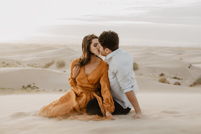 engaged couple sitting in sand dunes kissing