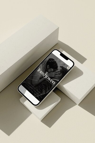 A smartphone displaying a wedding photography website, featuring a black-and-white image of a couple kissing. The text reads, 'Tom Bowen Photography, A Birmingham Wedding Photographer That Helps You Have Your Day, Your Way.' The phone is placed on beige geometric blocks.