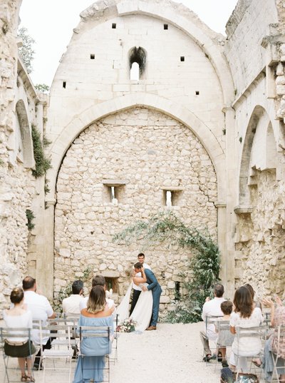 bride and groom kissing in front of a stone wall as crowd watches