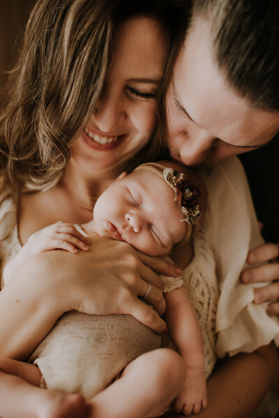 A mom holding her newborn baby to her chest while dad bends over her shoulder and kisses the baby on top of the in a boho photography studio in fort worth, texas