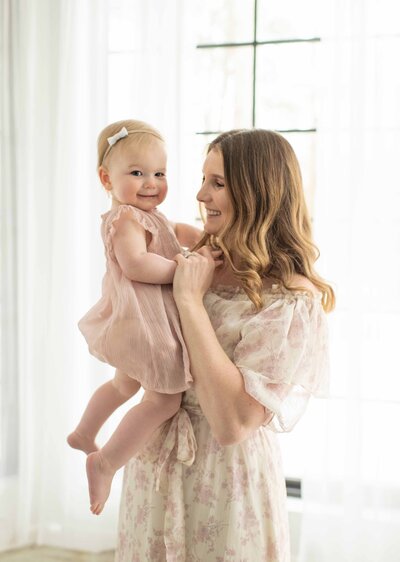 Mom in a flowing floral dress lifts her smiling baby near a bright window. A joyful motherhood session by Moments in Light Photography.