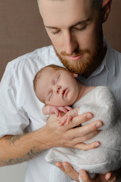 Close-up portrait of a father tenderly cradling his newborn against his chest, highlighting quiet connection and love. Photographed by Kaitlyn Dawn Photography in the Inland Empire.
