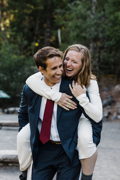 A blond bride wearing a strapless white gown holds it up while walking through the muddy trail in her hiking boots while her groom in glasses holds her waist and carries a dried floral bouquet while looking at his bride as they hike down the Elk Flats Trail near Manzanita Oregon for their adventure elopement on the coast. | Erica Swantek Photography