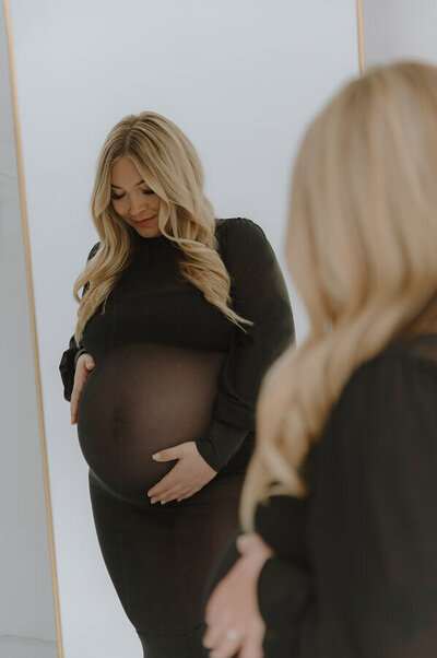 Wearing a sheet black dress that shows off her belly, a woman holds her pregnant belly. She's standing inside of a white studio.