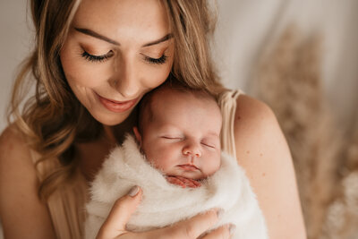 Photo of a sleeping newborn baby held facing outwards towards the camera by her mother who is cuddled in close to her and smiling during a photoshoot