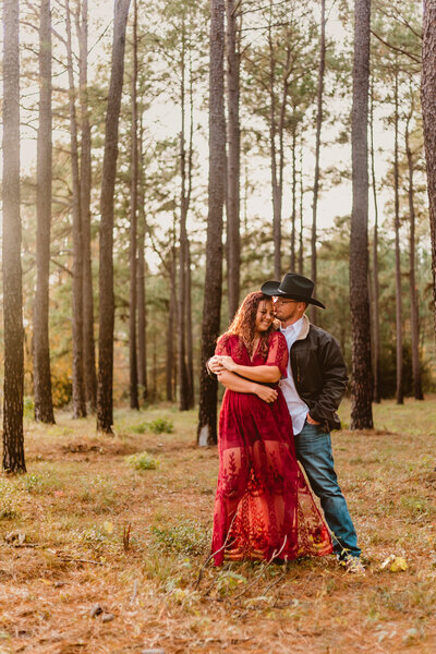 Couple embracing in a pine forest during their engagement session. The woman is wearing a flowing red dress while the man is wearing a hat and denim, sharing a romantic moment surrounded by tall trees. Captured by ShutterUp Photography.