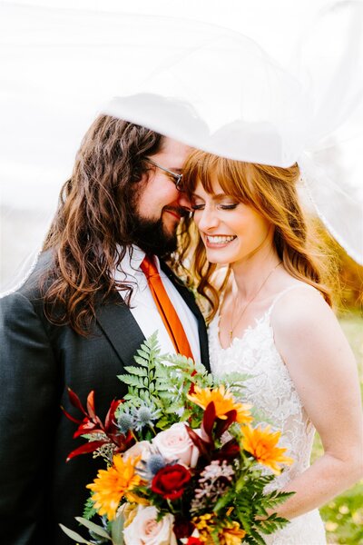A bride and groom share a close, intimate moment under her flowing white veil. The bride, smiling with her eyes closed, has long wavy hair and wears a lace gown. The groom, in a dark suit with a long orange tie, nestles his face next to hers. They both hold a bouquet with a mix of autumn-colored flowers, including red roses and yellow sunflowers.