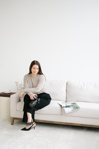 Ephe sits on a white couch and crosses her legs while holding her camera down by her side. 