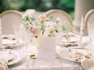 An elegantly set table with a floral centerpiece, crystal glassware, white plates, gold utensils, and white twisted taper candles curated by a top Calgary wedding planner. A card reading "three" is placed at the center.