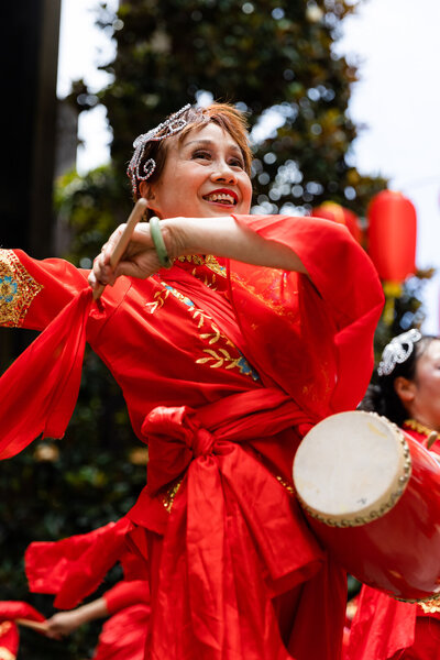 Event photography at Top Ryde City in Sydney showing a Chinese lady dancing a traditional dance and wearing a bright red traditional custume. It is a Lunar New Year celebration and she is smiling and happy whilst playing a drum
