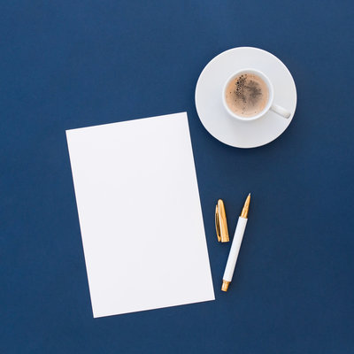 Flat lay of a white sheet of paper, a gold and white pen, and a cup of coffee on a navy blue background, symbolizing creativity, focus, and productivity in a clean and elegant workspace