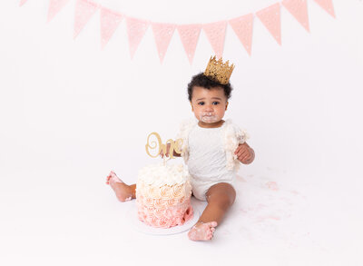 Baby girl is sitting against a white backdrop wearing a white lace onesie and a gold crown on her head. Between her legs is a blush ombre rosette cake. Baby girl has cake on her face, hands, and feet. Captured by top NYC and Brooklyn Newborn Photographer Rochel Konik.