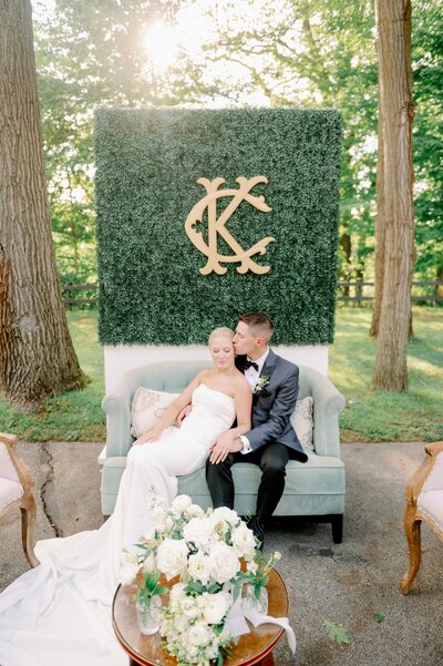 groom kissing bride as they sit on a sofa in front of a greenery wall with their initials on it