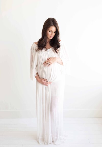 Pregnant mother in a flowing white gown cradles her belly in a bright, minimalist studio. A timeless maternity session by Moments in Light Photography.