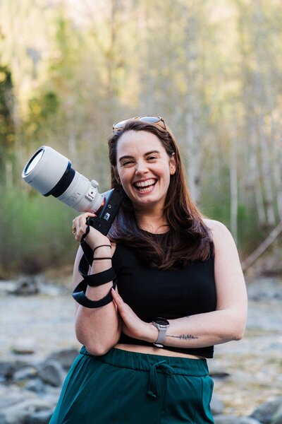 Alexandria Odekirk, the elopement photographer at Sage Trails, smiling while holding a camera along rocky river side in Washington