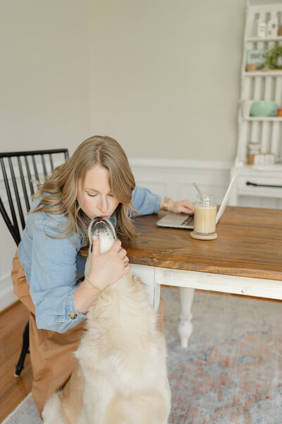 Woman bending over to the side of the table to kiss an english-cream golden retriever on the nose
