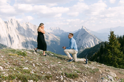 Groom on one knee proposing on a mountain top in Kananaskis Country during the summer after a hike with the bride saying yes captured as a romantic proposal moment