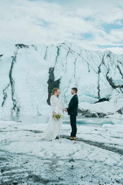 A man and woman hold hands while wearing wedding clothing on a glacier in Alaska