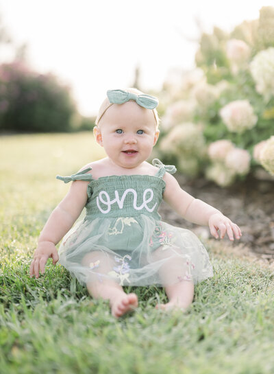 Smiling baby sitting on grass with pink hydrangeas in the background during an outdoor milestone session.