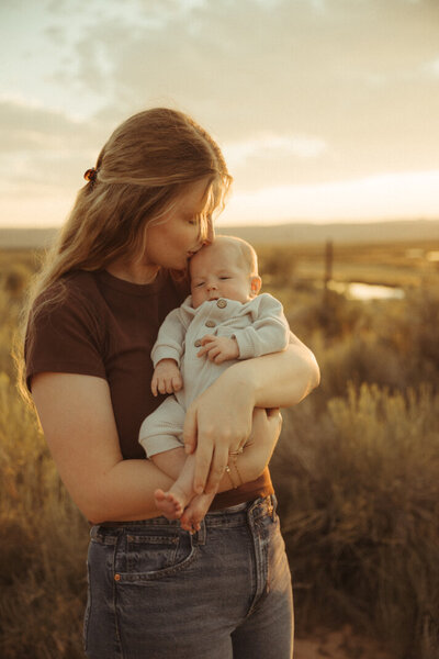 A precious portrait of Hannah from Hannah Grace Photo holding her young baby outside in Torrey, Utah. She is kissing her baby on the head.
