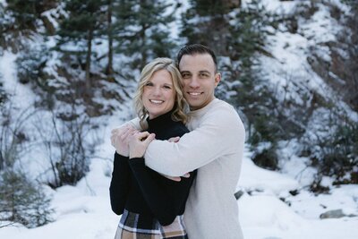 A man stands behind a woman wrapping his arms around her chest as they both smile at the camera.