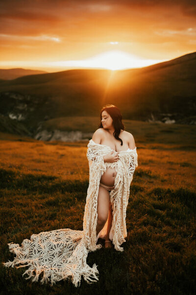 Pregnant mother draped in an antique blanket with the sun setting over the mountainsbehind her.