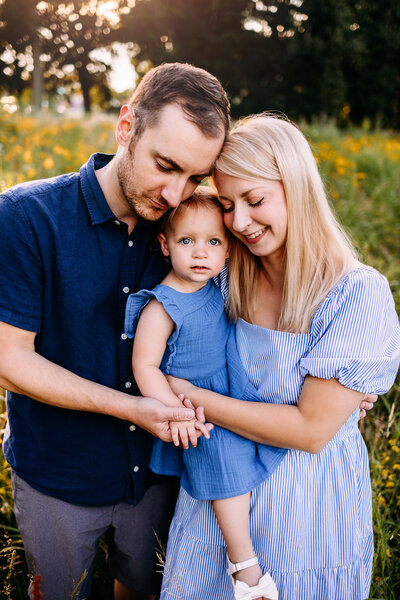 Mother and father snuggle cheek to cheek with their baby girl amongst a field of yellow flowers in Erie