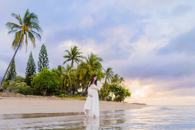 Pregnant woman holds her belly under palm trees and pastel skies in Hawaii